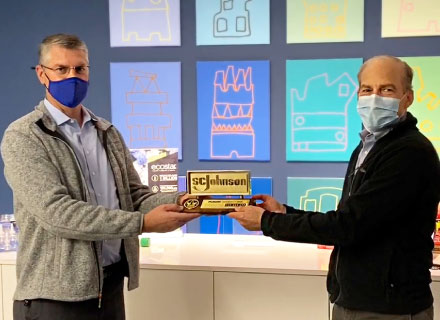 Two masked men present a branded cake or award box in front of colorful display panels