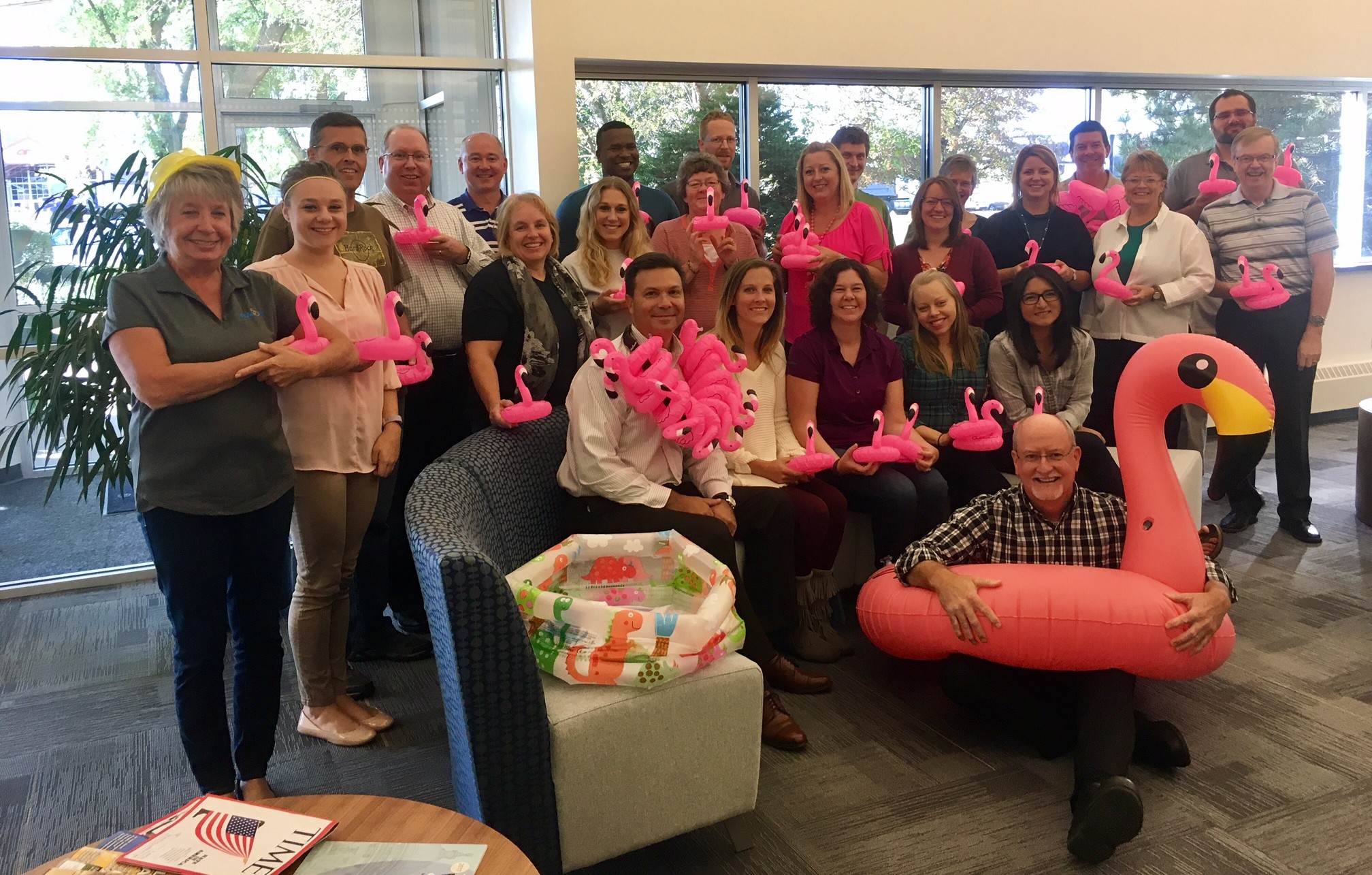 A group of coworkers pose indoors with pink flamingo decorations and a large inflatable flamingo