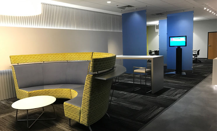 Modern office lounge with a curved yellow sofa, coffee table, high-top desk, and digital display