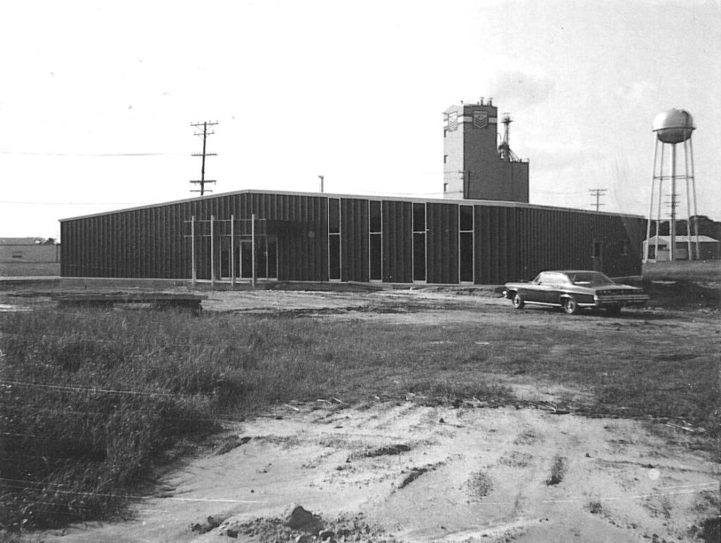 Low industrial building with a grain elevator, water tower, utility poles, and a vintage car